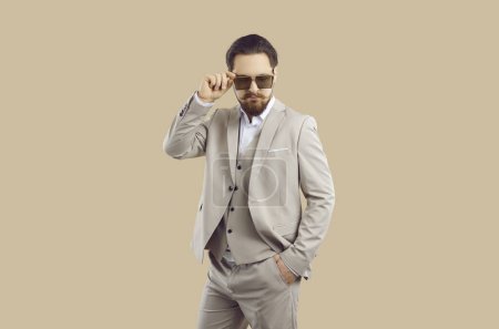 Male model in formal suit posing in studio. Attractive young man in modern elegant beige vest suit and cool sunglasses standing isolated on beige color background. Male fashion and style concept