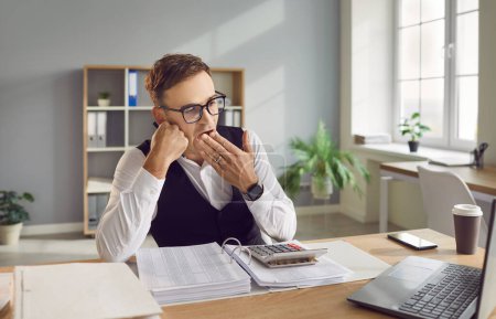 Photo for Tired, sleepy and bored financial accountant yawning at work. Man who didnt have enough sleep at night sitting at office desk with paperwork, laptop and coffee, yawning and covering mouth with hand - Royalty Free Image