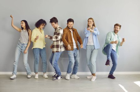 Six carefree cheerful young people dancing and having fun while meeting at home. Male and female best friends dressed in casual clothes are dancing standing in row against gray wall background.