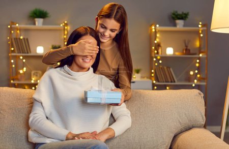 Photo for Positive daughter surprises mother at home, closing eyes and presenting a gift in a box to celebrate a holiday, Mothers day, Women day or special day. The family shares a happy moment together. - Royalty Free Image