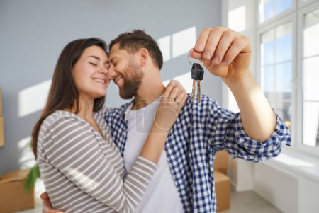 Photo for Happy young couple holding a key in hands standing in the living room at home hugging enjoying real estate or car purchase, smiling and celebrating moving day. Relocating, mortgage concept. - Royalty Free Image
