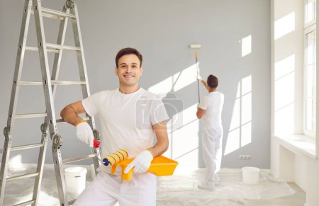 Photo for Male workers doing interior renovation in apartment. Portrait of happy smiling confident adult man wall painter in white uniform holding paint roller and tray standing by step ladder looking at camera - Royalty Free Image