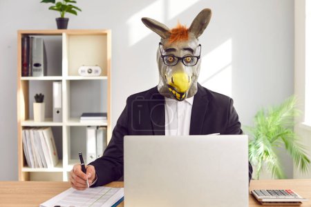 Corporate financial accountant in funny animal mask working in office. Man employee in unusual, bizarre, silly, humorous, wacky rubber donkey mask and eyeglasses sitting at desk with laptop computer