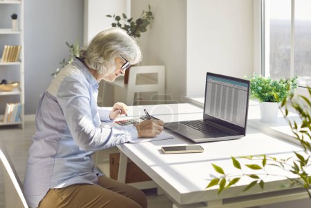 Concentrated serious senior accountant woman sitting on workplace at the desk working on laptop with charts making notes calculating finances, taxes or budget. Pension calculation concept