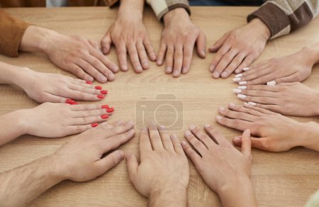 Téléchargez les photos : Groupe de jeunes ayant une réunion, assis en cercle autour d'une table et tenant leurs mains ensemble sur sa surface en bois. Équipe, travail d'équipe, communauté, entreprise, concept de connexion - en image libre de droit