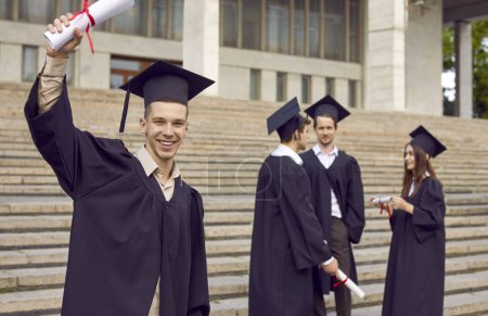 Photo for Happy male student having fun on graduation day. Portrait of joyful young man in academic hat and gown standing outside college or university building, showing diploma, looking at camera and smiling - Royalty Free Image