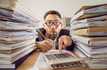 Overworked business man in suit and funny glasses working at the desk on his workplace at office with a pile of folders and a stack of papers. Tired accountant making calculations analyzing data.