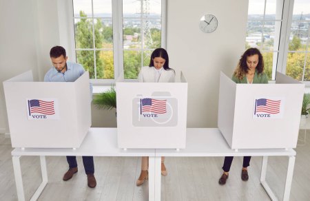 Group of young confident American citizens voters standing in a row at vote center with USA flags in voting booth. People voting at polling station on election day. Democracy concept.