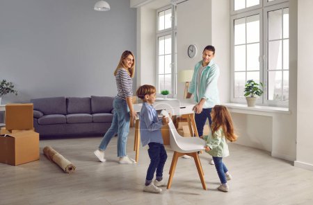 Photo for Happy family couple with two kids boy and girl arranging furniture table and chairs in a new apartment on moving day with unpacked boxes in background. Relocating, real estate, mortgage concept. - Royalty Free Image