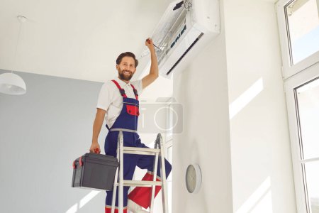 Photo for Cheerful air conditioner technician, dedicated to maintaining and repairing cooling systems. Serviceman conducts maintenance, repair or install the unit while standing on a ladder in the room. - Royalty Free Image