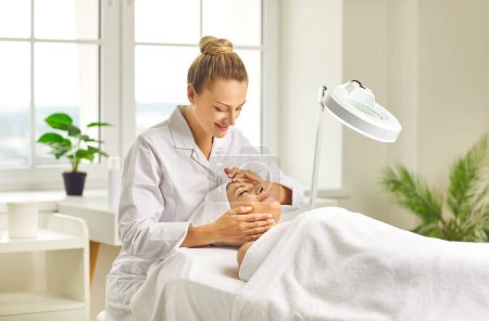 Photo for Young woman cosmetologist or dermatologist checking skin condition to a smiling girl client with special lamp in beauty spa salon. Facial massage, skin care treatment and cosmetology concept. - Royalty Free Image