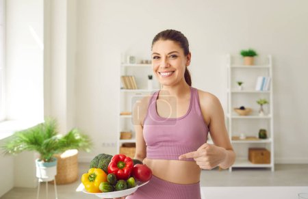 Photo for Young woman keeps fit by working out and eating healthy food. Portrait of happy beautiful girl in pink sports top smiling and pointing at plate with colorful raw vegetables. Fitness concept - Royalty Free Image