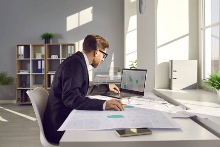 Photo for Professional cadastral surveyor in business suit sitting at table in modern office, using laptop computer, working with city structure design plan, looking at map, studying plot numbers and boundaries - Royalty Free Image