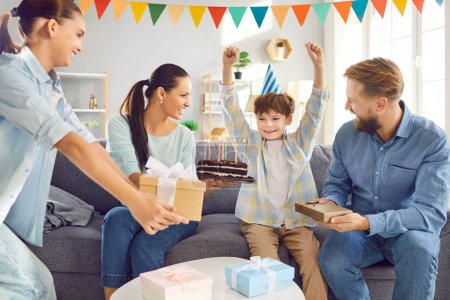 Photo for Happy Caucasian family surprises son with gifts during birthday party. He rejoices, sitting on the sofa in the living room at home, celebrating special day with happiness and the love of family. - Royalty Free Image