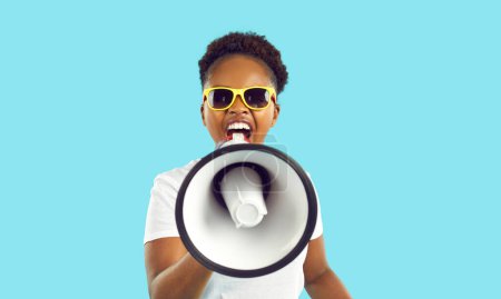 Fröhliche verrückte afrikanisch-amerikanische Frau mit Lautsprecher in der Hand, die laute Werbung auf hellblauem Hintergrund macht. Junge Frau in weißem T-Shirt und Sonnenbrille schreit laut über verrückte Rabatte.