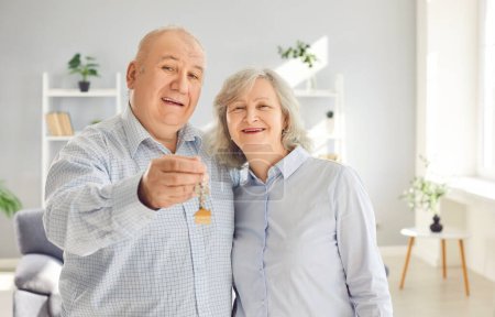 Foto de Feliz pareja de ancianos sosteniendo una llave en las manos de pie en la sala de estar en casa mirando a la cámara disfrutando de la compra de bienes raíces, sonriendo y celebrando el día en movimiento. Reubicación, concepto de hipoteca. - Imagen libre de derechos