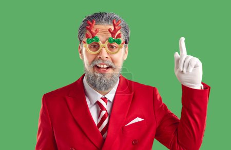 Téléchargez les photos : Homme barbu aux cheveux gris dans des lunettes de Noël drôles avec des bois, costume rouge ayant une bonne idée pointant index vers le haut sur fond vert et en regardant la caméra. Noël, Nouvel An, bannière de vacances d'hiver. - en image libre de droit