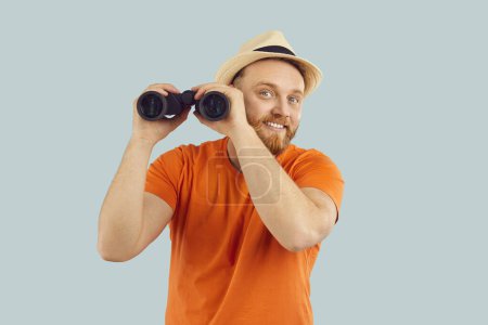 Curious Caucasian bearded man tourist smiles at the camera holding binoculars in hands, expressing contentment and curiosity, against a blue background in a portrait of discovery and search.