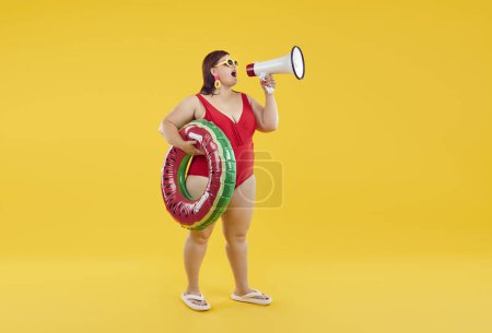 Photo for Full body photo of a funny fat overweight woman wearing red swimsuit holding rubber ring and speak in mouthpiece isolated on a studio yellow background. Summer holiday trip and vacation concept. - Royalty Free Image