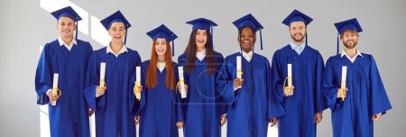 Foto de Cartel con retrato grupal de felices estudiantes universitarios multiétnicos de raza mixta sonrientes y felices con diplomas en gorras de graduación azules y vestidos de pie juntos sobre fondo de pared gris claro - Imagen libre de derechos