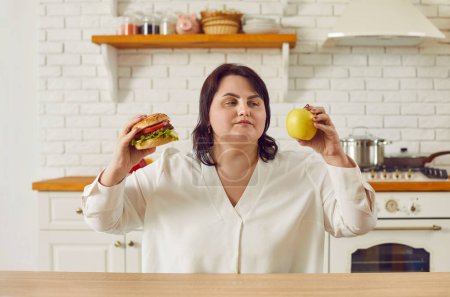 Photo for Overweight, plump brunette woman choosing between healthy and unhealthy eating. Puzzled plus size woman choosing between burger and apple while sitting at table in kitchen. Healthy and unhealthy - Royalty Free Image
