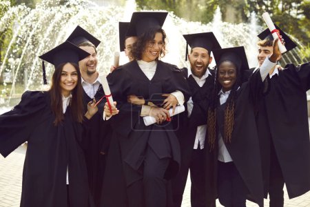 Portrait of a group of smiling happy multiracial international graduates students having fun in a university graduate gown and holding diploma outdoor. Education and graduation concept.
