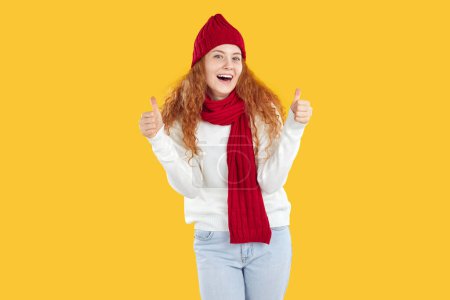 Téléchargez les photos : Joyeux amical rousse bouclé souriant fille positive en chapeau rouge, écharpe, pull blanc et jeans montrant pouces vers le haut en regardant la caméra sur fond jaune. Bannière pour la publicité, marketing, ventes. - en image libre de droit