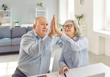 Senior couple sitting at the desk and giving high five each other calculating finances, taxes and budget at home. Elderly retired man and woman planning income and profit on pension.