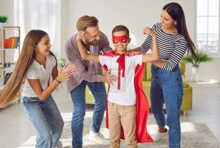 Photo for Little child is ready to protect people and save world. Cheerful kid in superhero clothes having fun with family. Happy smiling boy wearing super hero suit shows biceps muscles to mum, dad and sister - Royalty Free Image