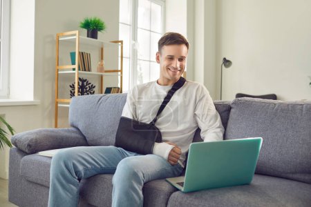Hombre sonriente con un brazo roto en la lesión consecuencia, envuelto en un yeso protector y vendaje, muestra resistencia como él cómodamente a pesar del trauma utiliza un ordenador portátil en el sofá de la sala de estar en casa.