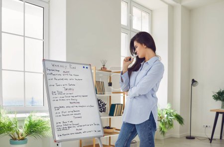 Portrait of a young confident business woman standing in office or at home in front of white board with daily plans and tasks. Girl looking at work schedule planning appointment for week.