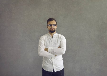 Studio portrait of guy in funny sunglasses. Serious young man in white shirt and thug life meme glasses standing arms crossed isolated on grey background
