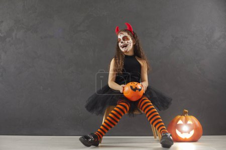 Téléchargez les photos : Joyeux enfant fille avec maquillage festif en costume noir de carnaval du diable avec des cornes assis dans le studio sur fond gris avec espace de copie tenant orange citrouille d'Halloween et riant. - en image libre de droit