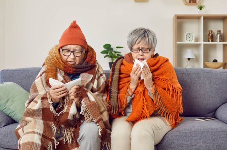 Téléchargez les photos : Grippe comme symptômes. Le couple âgé congelé est malade à la maison pendant le rhume d'automne et souffre de rhume. Senios malades, chaudement habillés et enveloppés dans des couvertures, essuient le nez avec des serviettes tout en étant assis sur le canapé. - en image libre de droit