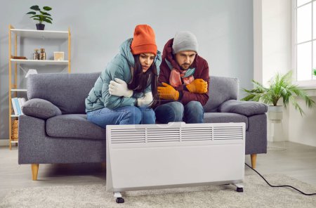 Porträt eines jungen, gefrorenen Paares, das frustriert in winterlicher Oberbekleidung und Mützen zu Hause auf dem Sofa im Wohnzimmer sitzt und versucht, sich neben der Elektroheizung warm zu halten. Heizungsprobleme.