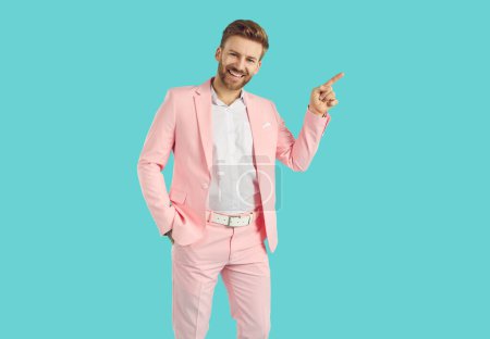 Photo for Happy man pointing to side. Handsome young man in pink suit standing with hand in pocket isolated on blue background, showing something, advertising fashion sale, smiling and pointing his finger away - Royalty Free Image
