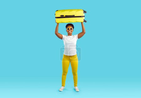 Foto de Retrato completo de una mujer feliz afroamericana con una maleta amarilla sobre su cabeza sonriendo sobre fondo turquesa. Lleva vaqueros amarillos y camiseta blanca. Viajes, turismo, viajes - Imagen libre de derechos