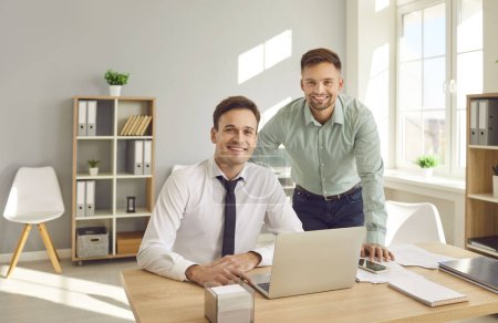 Foto de Portrait of a two happy smiling confident business people wearing shirts looking cheerful at camera at the workplace sitting at the desk. Company employees and male coworkers working in office. - Imagen libre de derechos