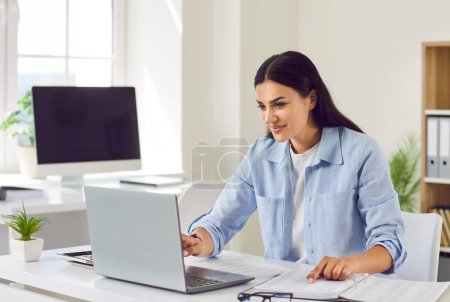 Photo for Positive woman works with papers and a laptop at home office or a traditional workspace. Her commitment to productivity seamlessly blends online technology and traditional paperwork. - Royalty Free Image