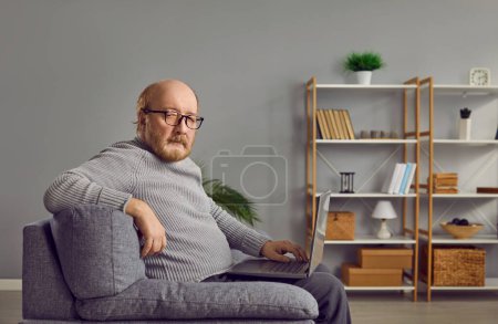 Homme âgé assis sur un canapé avec un ordinateur portable et souriant à la caméra. Homme âgé sérieux dans les lunettes de vue portant pull tricoté gris tenant ordinateur portable sur ses genoux tout en étant assis sur le canapé à la maison