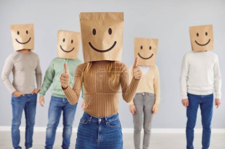 Photo for Customer with smiley bag on head feels happy, leaves positive review, gives thumbs up. Portrait of casual woman in shopping bag with cheerful face emoticon in studio with group of people in background - Royalty Free Image