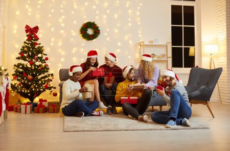 Photo for Christmas mood. Group of young people exchange gifts and open gift boxes sitting by Christmas tree. Joyful and surprised multiracial people in Santa hats sit on sofa and on floor in cozy room at home. - Royalty Free Image