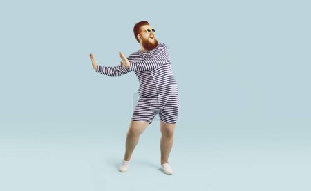 Photo for Funny fat man in striped pyjama isolated on blue studio background dancing. Smiling overweight redhead guy in sunglasses have fun make dancer moves. Positive lifestyle and entertainment. - Royalty Free Image