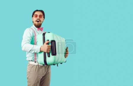 Photo for Cheerful man in shirt, pink glasses, bowtie and classic pants with suspenders standing on turquoise copy space background, holding his suitcase and looking at camera. Holiday, traveling, trip concept - Royalty Free Image