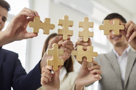 Photo for Happy smiling group of business people assembling wooden puzzle standing in office. Company employees join jigsaw pieces finding solution. Cooperation, teamwork, help and support concept. - Royalty Free Image