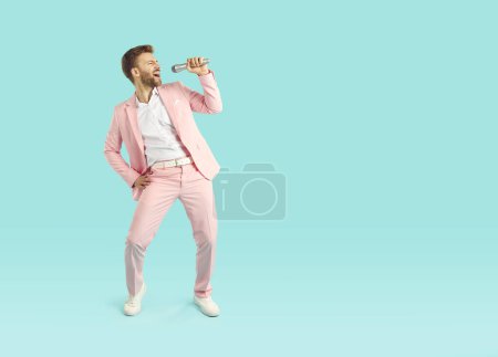 Foto de Joven hombre alegre y elegante en traje rosa cantando canción en el micrófono aislado en el fondo azul del estudio. Retrato emocional del cantante expresivo y divertido en el club de karaoke. Concepto de vida. - Imagen libre de derechos
