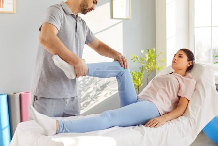 Photo for Physiotherapist treats leg injury and helps patient perform exercises for lower limbs. Female patient lies on medical couch in rehabilitation room and male doctor holds her foot and stretches her leg. - Royalty Free Image