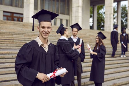 Photo for Portrait of joyful smiling male graduate student in a university gown looking cheerful at camera holding diploma in hands. Happy confident guy standing outdoor with classmates in background. - Royalty Free Image