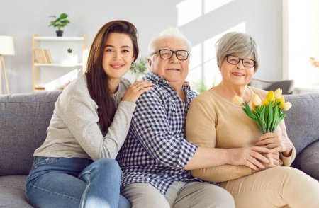 Foto de Retrato de una joven mujer alegre sentada en el sofá con sus viejos padres mayores en la sala de estar en casa felicitando a su madre madura feliz con el cumpleaños o el día de las madres presentando ramo de tulipanes - Imagen libre de derechos