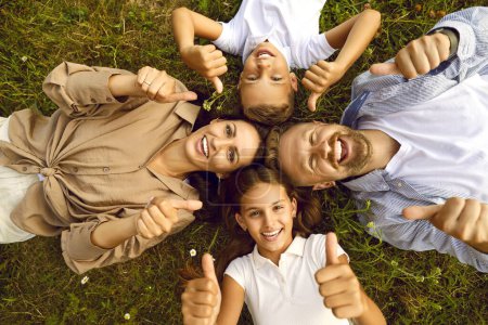 Photo for Top view of happy young family of four lying on green grass and showing thumb up sign in summer park. Smiling parents having fun with kids boy and girl in nature enjoying time together. - Royalty Free Image